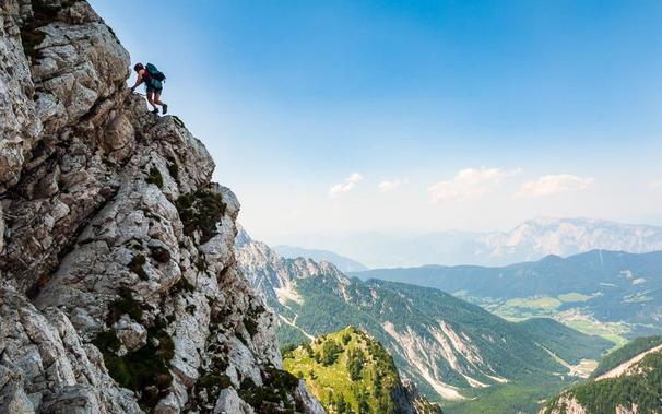 A silhouetted figure, small in the frame, is outlined against blue sky on the edge of a steep rock buttress. A green valley lies more than a thousand metres below, with more mountains, though less steep, seen beyond, slightly softened by haze.