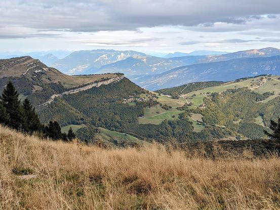 A wide, slightly high-angle shot captures a vast mountainous landscape under an overcast sky. In the foreground, dry, golden-brown grasses dominate, slightly blurred. Beyond this, a series of rolling hills and mountains stretch into the distance. The closest mountains are covered in patches of green vegetation and darker evergreen trees. A distinctive, long ridge with a visible path or road running along its top cuts across the mid-ground. Further back, layers of progressively lighter blue-grey mountains fade into the horizon, some with hints of snow on their higher peaks. The sky is a uniform grey, suggesting a cloudy day. The overall impression is one of expansive natural beauty and a sense of remoteness.
