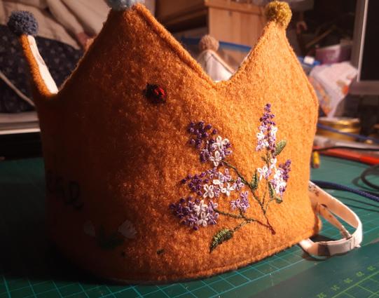 Detailaufnahme einer Geburtstagskrone aus orangenem Wollwalk. Lilane Fliederblüten und ein kleiner Marienkäfer sind auf die Krone gestickt. Kleine Bommeln zieren die Spitzen der Krone. Ein Band liegt seitlich an der Krone.