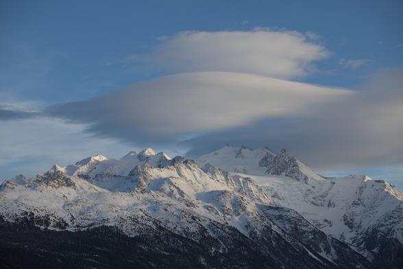 Berglandschaft im Wallis: Blauer Himmel mit Föhnfischen über den Berggipfeln des Mischabel-Massivs. Die Berge sind noch eingeschneit, die Baumgrenze unterhalb ist dunkel-braun.
---
Mountain landscape in Valais: Blue skies with cumulus lenticularis clouds over the peaks of the Mischabel massif. The mountains are still covered in snow, the tree line below is dark brown.