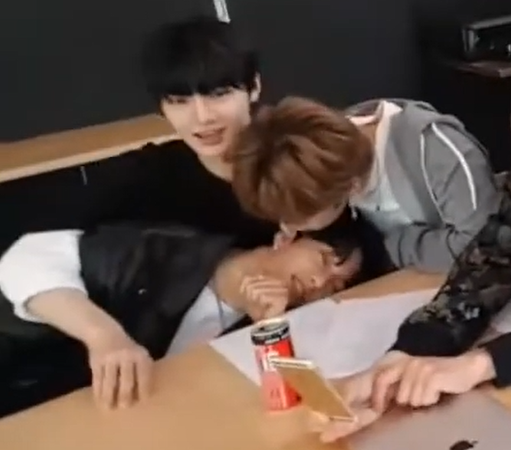 Hyunjin leans over almost sideways onto I.N's lap, while Lee Know bends over Hyunjin and is about to kiss the side of his face.