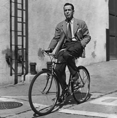 Humphrey Bogart con chaqueta y corbata montando una bici en un estudio de Hollywood, posiblemente durante la grabación de la película Sabrina en los años 50