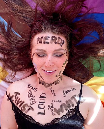 Bild von Lili von Oben, ab Brust aufwärts. Sie liegt, nur bekleidetet mit einem schwarzen BH, auf einer Progress Pride Flagge. Die rötlich braunen Haare gehen zu allen Seiten weg. Sie trägt silbernen Lidschatten und einen dünnen dramatischen schwarzen Lidstrich mit Wing. Sie hat leichte Sommerprossen und zwei klar definierte gerade Augenbrauen, die Rechte davon mit einem tiefen Cut. Auf ihren Körper sind mit schwarzer Farbe in verschiedenen Schriftarten viele Begriffe gemalt, die sie komplett bedecken. Farbe hat nur das kleine "trans" auf einer trans Pride Flagge auf der rechten Wange. Die Worte sind: Activist, caring, vegan, left, loud, artist, feminist, cringe, white, women, nerd, gamer, aunt, daughter, wife, sister.