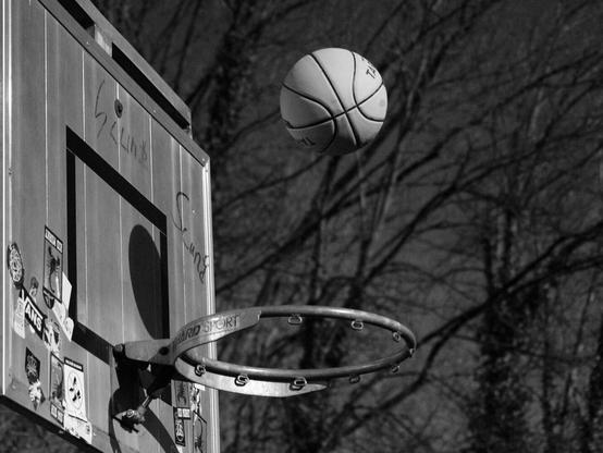 A monochrome image features a basketball in mid-air, shot toward a basketball hoop. The hoop is mounted on a weathered metal backboard adorned with faded stickers and graffiti, adding character. The background is blurred, showing bare trees. The scene conveys a sense of urban outdoor play, slightly worn but still inviting.