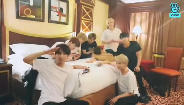 skz in a hotel room. Seungmin is bend over the bed with his head sideways on a cushion and one arm stretched out in front of himself. He looks relaxed and comfortable. Chan's standing behind him and Changbin sits next to him on the bed, leaning towards him.