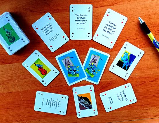 Auf einem Tisch liegen mehrere Spielkarten mit den üblichen Zahlen und Motiven von Skat-Karten, aber ergänzt um Abbildungen von gemalten musizierenden Kätzchen und passenden Sprüchen.