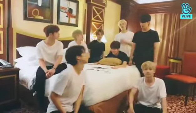 skz in a hotel room. Seungmin is bent over the edge of the bed and doesn't need to hug the cushion any more. He has both arms extended in front of himself with his palms flat on the bed. Chan's standing very close behind him.