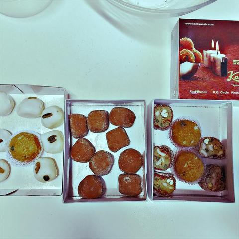 This picture shows a collection of sweet treats displayed on a white surface, with some items still in their packaging, and what appears to be traditional Indian desserts such as ladoos and halwa. The mood or intention seems to be that of showcasing the variety and presentation of these desserts.