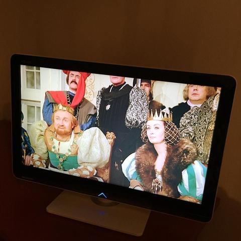 This picture shows a television screen displaying a scene from a movie or TV show featuring individuals dressed in elaborate period costumes. The main focus is on two central figures who appear to be royals, possibly of a fictional or historical setting, dressed in ornate attire with the male character wearing a crown and the female character adorned in a gown with what seems to be a necklace made from jewelry. Both characters have expressions of seriousness on their faces. The background shows other individuals, also dressed in period costumes, suggesting they are part of the same social gathering or event. The overall mood conveyed by the image suggests a setting of historical importance, possibly a royal court or assembly.