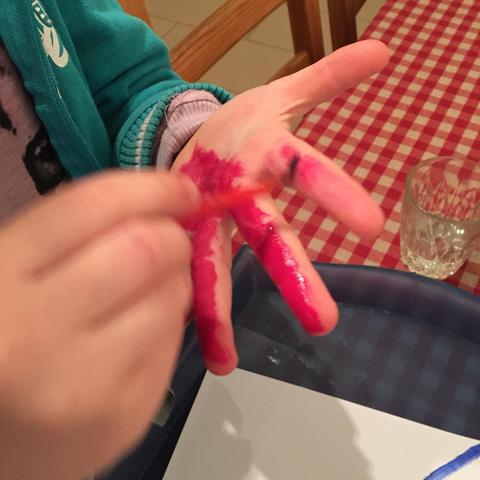 This picture shows a young child sitting at a dining table, engaging with an activity involving red paint on their fingers. The child is attentively examining their hand, which has dried paint on it. They appear to be using the paint to create something on a piece of paper placed before them, possibly a craft or art project. The setting suggests a casual, domestic environment, with a glimpse of a cup and a tablecloth in the background. There are no visible texts on the image.