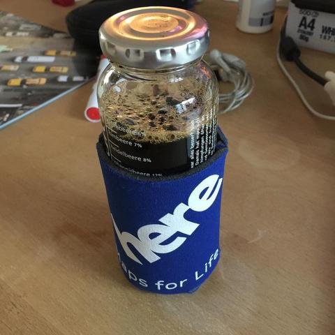 This picture shows a jar filled with a black liquid, placed inside a blue cup cozy, which is sitting on a table next to some papers. The setting suggests someone might be working or studying.
