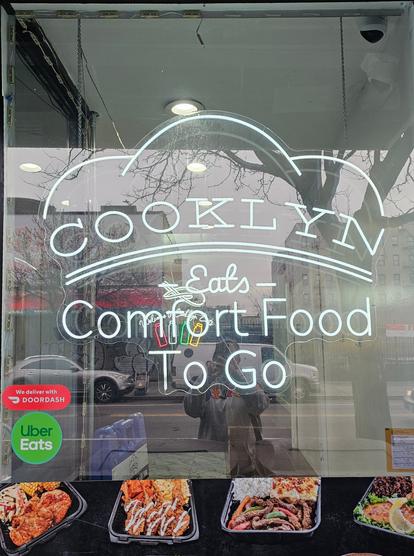 Photo of a storefront window tgat reads 'COOKLYN Eats Comfort Food To Go" in white neon tubing. In tge background there are 4 neon tube bottle shapes in red, white, green, yellow. Thd wibdow has a bright red paper sticker foe Door dash and a green one foe uber eats