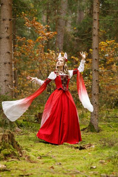 A woman dressed in a long, red, fantasy gown is standing in the woods. She has white fox ears (on top of her head) and wears a flower headpiece. The goen has long sleeves.