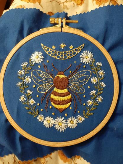 Broderie dans son cercle de bois. Le tissus de base est bleu, le motif central est un bourdon, autour de lui des fleurs, tiges, feuilles, et au dessus une lune et 3 étoiles