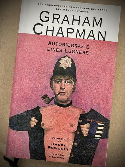 The image features a book cover for Graham Chapman's autobiography titled "Autobiografie eines Lügners." The cover displays an illustration of a man in a police uniform, smoking a pipe, and exposing his chest.