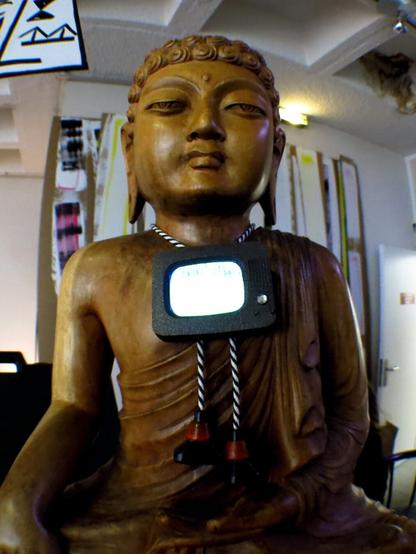 La statue de Bouddha en bois, en situation dans le lounge de Videoformes 2017, portant le Vijou 'VintageTV' en forme de télé des années 60 et intégrant un écran vidéo, monté en bolo-tie avec des embouts de cordes en forme de tube jouant la ligne blanche constante de Zen TV.