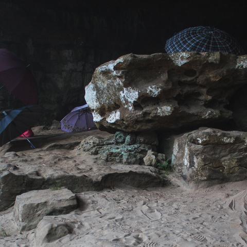 A photo of an ancient cave with various weathered rock formations and sand. Several opened umbrellas have been placed on the rocks and there are many footprints in the sand.