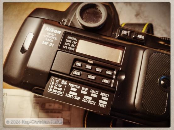 Rückansicht einer analogen Nikon F801s Kleinbild-Spiegelreflexkamera mit der Datenrückwand MF-21, deren Deckel über den Bedienelementen aufgeklappt ist und so den Blick auf diverse kleine, kompliziert beschriftete Bedienknöpfe freigibt.