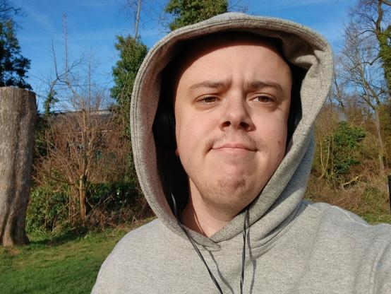 A white transmasc wearing a grey hoodie stood in a park