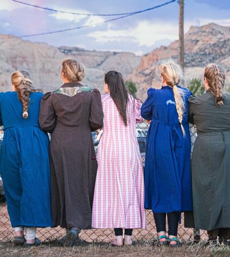Photograph of five women, their back to the camera as they gaze at a mountain scene. The women are dressed in the old-fashioned outfits worn by adherents of fundamental Mormonism.