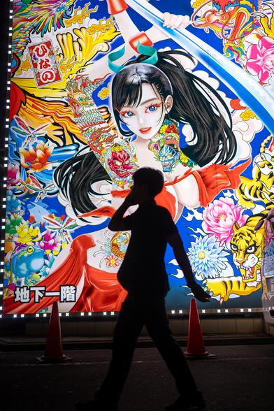 Silhouette of a man walking in front a colourful illuminated poster in a dark street in Tokyo at night. The poster features a manga-style girl with dark hair and impressive breast who seems to be looking in the direction of the viewer. She is surrounded by flowers and a tiger.