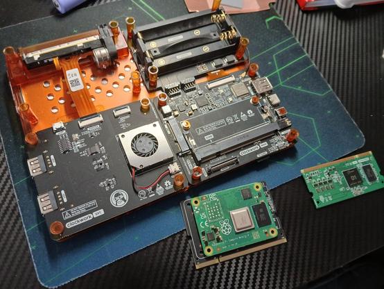 Das Mainboard des DevTerms liegt auf einem Tisch in Carbon-Optik, vor dem DevTerm liegen links das CM4 mit seinem Adapterboard und rechts das "alte" Allwinner D1 SoC