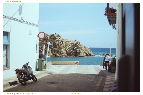 A photo of a sunny street with a sea at its end, and a brown rock formation sticking out. There's a moped in the bottom left, and a 'no traffic' sign that's grabbing attention. 
Sunny, hot atmosphere.