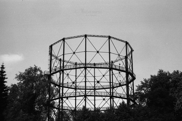 Quite dark black and white film photo of an old gasometer or rather its steel framework, surrounded by trees