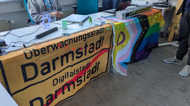 Ein langer Tisch mit Laptops, LEDs und 3d gedruckten Dingen.
Davor hängt eine Prideflagge uns ein Ortsschild zur Überwachungsstadt Darmstadt