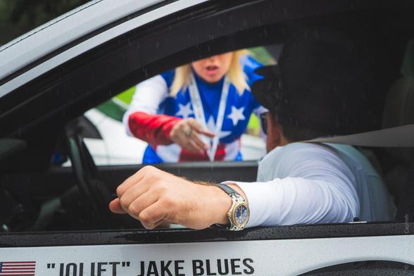 A man dressed as "Joliet" Jake Blues from The Blues Brothers leans over to talk to a woman on the other side of the car, during the Cannonball Run in Blarney, Cork, Ireland, 2018. His arm rests on the window frame, wearing a silver watch, while he interacts with a person dressed in a red, white, and blue costume. The car door displays the text: "JOLIET" JAKE BLUES.