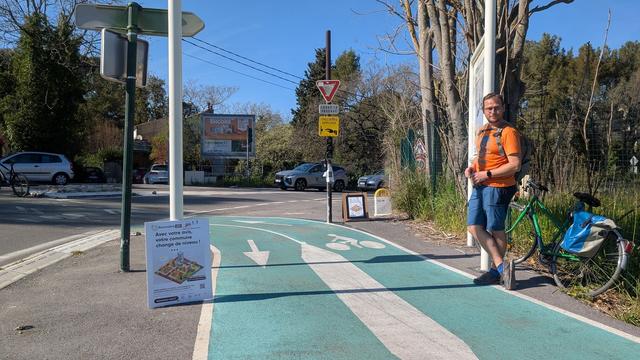 Photo: un homme blanc attends sur la piste du littoral entre Toulon et Le Pradet, à côté d'un feu. Deux affiches pour le baromètre vélo sont posées sur les deux côtés de la piste.