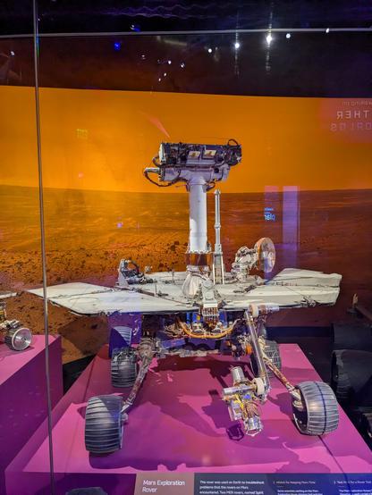 The Mars Exploration Rover 'surface system testbed' rover in a glass frontedes display. The rover is up on a magenta pedestal with a photograph taken by the Spirit Rover as the background.

A thin layer of dust from the simulant used during the testing campaign to free Spirit from Scamander Crater coats the rover's deck...hence the nickname, Dusty.