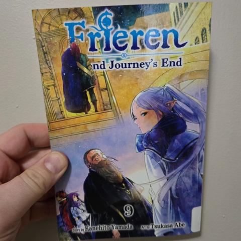 Photograph of a white person's hand holding Frieren: Beyond Journey's End, volume 9. Image shows the titular Frieren standing next to an older, bearded gentleman (Denken) with a white-blue background near the bottom half, a golden background and a colorful demon seen from behind in the upper half of the cover.