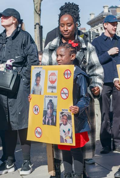 A woman stands with her young daughter who is holding a poster with photos of herself  showing the effects of topical steroid withdrawal on her skin.
