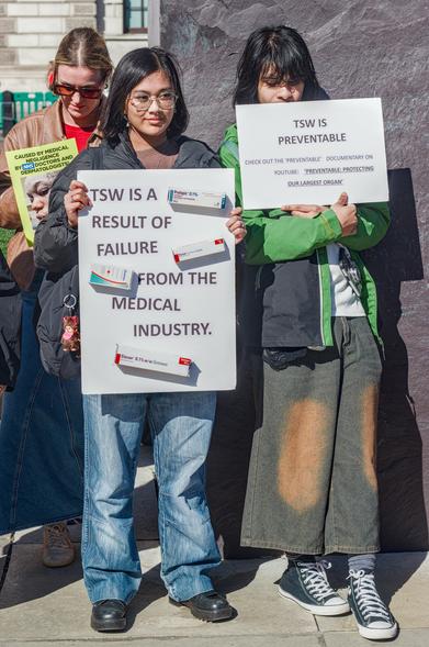 Two young women hold posters that read, 'TSW is a Result of Failure from the Medical Industry, and 'TSW is preventable.'