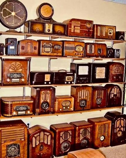 Das Bild zeigt eine Sammlung von alten Radios, die auf mehreren Regalen an einer Wand präsentiert werden. Die Radios stammen aus verschiedenen Jahrzehnten und haben überwiegend Gehäuse aus Holz mit kunstvollen Designs. Einige Modelle besitzen dekorative Lautsprecheröffnungen, während andere mit Drehknöpfen und Anzeigen ausgestattet sind. Ganz oben ist ein großer, runder Lautsprecher mit einem blumenartigen Muster zu sehen. Die Geräte sind in verschiedenen Größen und Formen arrangiert, darunter rechteckige, quadratische und abgerundete Designs. Die Wand hinter den Regalen ist beige, und die Regale bestehen aus Metallstreben mit Holzablagen. Die Sammlung vermittelt einen nostalgischen Eindruck und zeigt die Vielfalt des Radio-Designs aus der Vergangenheit.