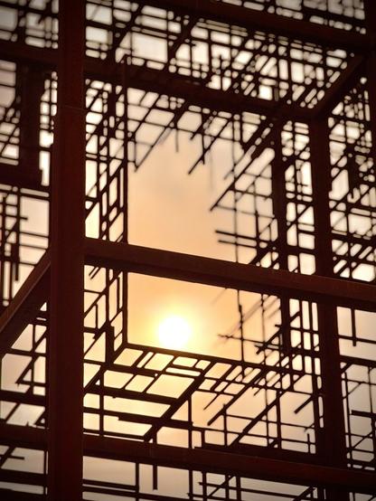 Numerous burgundy steel bars and beams intersect at 90-degree angles, as the sun shines through the semi-cut-out shape of a window within them.