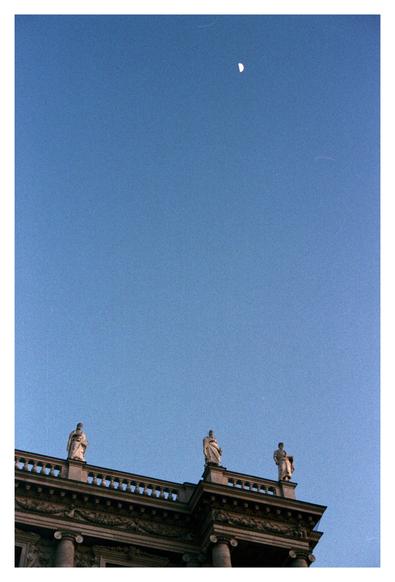 Color photograph of three parapet figures of the Natural History Museum Vienna, taken from Bellariastrasse at dusk with a deep blue sky and the crescent moon high above the figures.