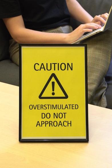 Framed sign "Caution! Overstimulated. Do not approach" is standing on a desk with a person sitting on a sofa behind it.