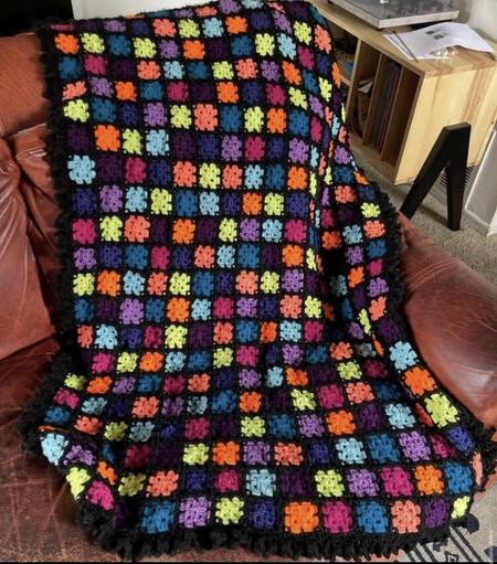 A colorful blanket on a red, leather couch. The blanket is made up of small granny squares in the colors dark pink, light purple, dark purple, light teal, dark teal, bright lime-ish green, orange, and dark blue. The squares are joined with a braided join in black yarn. There is a small black border around the blanket.