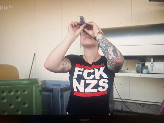 junge Winzerin prüft den Zuckergehalt von Weintrauben. Sie ist an den Armen tatöwiert und trägt ein schwarzes T-Shirt mit einem großen "FCK NZS"-Aufdruck in weiß der oben und unten umrahmt ist mit einem roten Linien