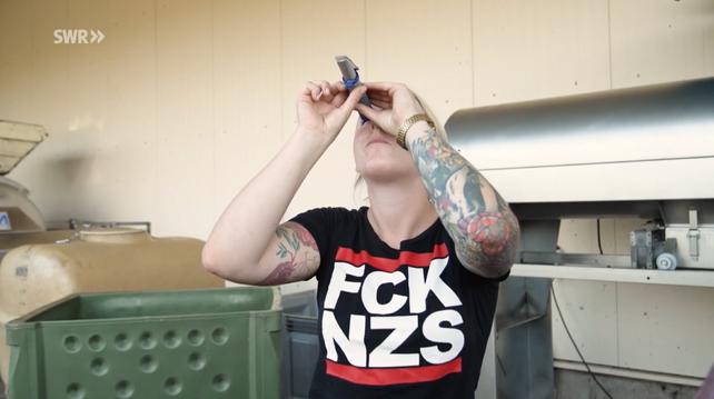 Junge Winzerin, die gerade den Zuckergehalt von Weintrauben misst mit schwarzen T-Shirt auf dem in großen weißen Buchstaben "FCK NZS"-steht, von oben und unten mit roten Linien umrahmt.