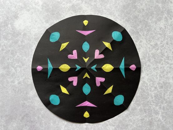 A paper stained glass style piece of art, made from a circle of round black paper with symmetrical shapes cut out. There is coloured tissue paper behind the shapes.