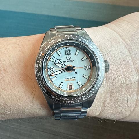 Watch with brushed stainless steel case with polished chamfer, brushed stainless steel H-link , world time bezel, pale blue dial with white numeric indices outlined in black at 12, 3 and 9, orange-on-brown date at 6:00 with date window outlined in orange, baton indices elsewhere, skeletonized black sword hands with white lume, orange GMT hand, arrow seconds hand, and 24-hour markings on the rehaut.