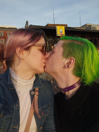 Two trans women kiss in warm lighting against a city backdrop. One has long, rose gold hair with dark roots tied back with turtleshell claw clip and is wearing glamorous sunglasses, silver earrings, a white ribbed turtleneck, a jean jacket, and the pink strap of a purse hangs across her body. The other trans woman has bob-length, electric green hair with an undercut, black earrings, a purple collar with a silver heart, and a chunky black sweater.