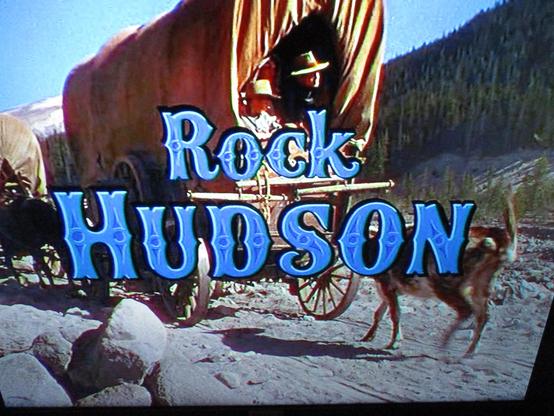 Générique du film de western Les Affameurs d'Anthony Mann (1952) avec à l'arrière-plan du nom de l'acteur Rock Hudson en grosses lettres bleu style cowboy, une caravane tirée par des chevaux et pleine de jeunes en chapeau passe près d'une forêt en altitude, par temps ensoleillé