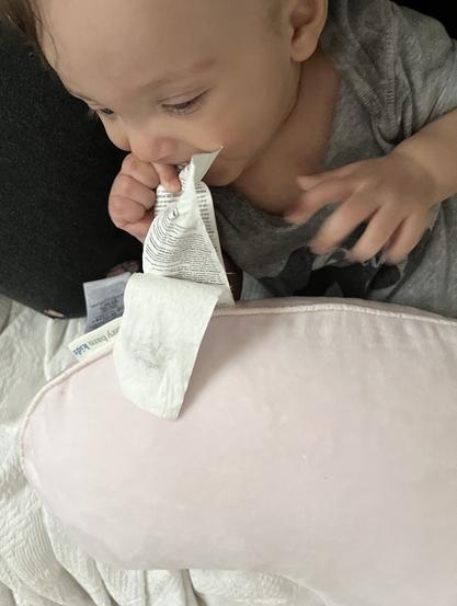A baby is sitting with a light pink pillow and is holding and exploring a large tag with their mouth.