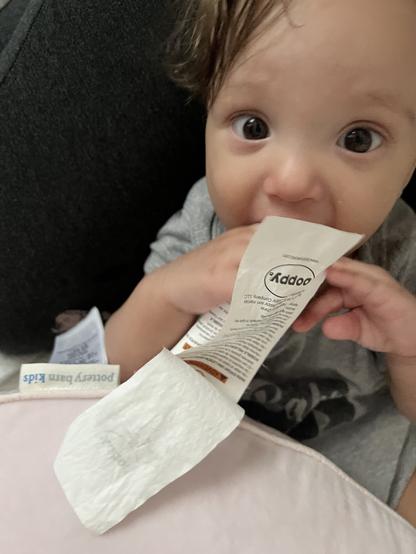 A baby is sitting and holding a tag, appearing to chew on it. They are looking directly into the camera with wide eyes.