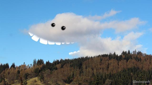 A photo of a wooded hillside with a blue sky above. There is a white fluffy cloud resting on the hilltop, which looks like a dog running to the left, mouth open, ears flapping behind in the wind. Well, it looks a little like that, and I've added some teeth and eyes to help.