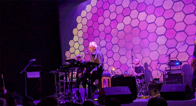 John Cale, lean and grey-haired, sits at a keyboard, centre stage, with a guitarist, drummer, and bass player (just out of shot) behind him. A purple and gold honeycomb of hexagons is projected behind the band. There's a fittingly sixties vibe to the scene.